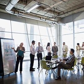 Standing woman explains charts to sitting man in office with other people standing and conversing.