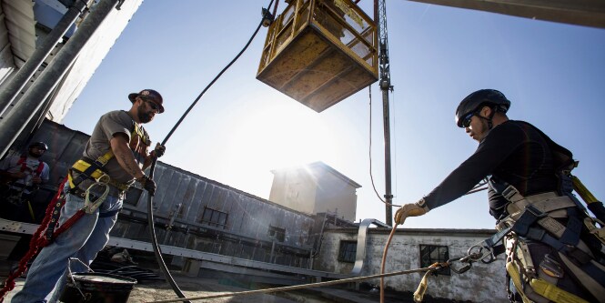 Image of two workers guiding hoses out of the way as a crane lowers a cage platform down to them
